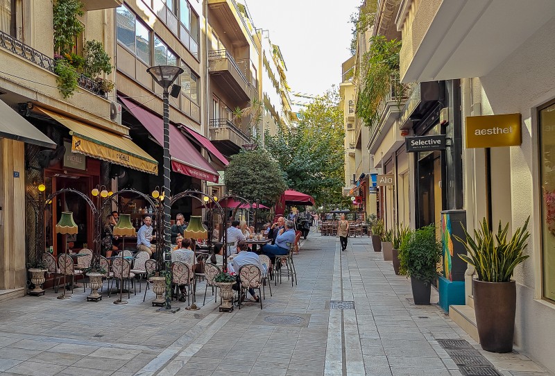 Outdoor café tables in a narrow Kolonaki street, Athens, with people enjoying coffee and boutique shops on both sides.