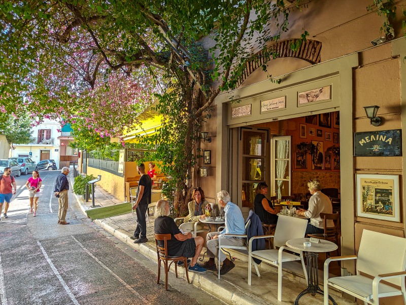 Lysiou Street in Plaka Athens with outdoor café tables, blooming flowers, and people enjoying coffee on a sunny morning, leading toward Anafiotika’s Cycladic-style houses.