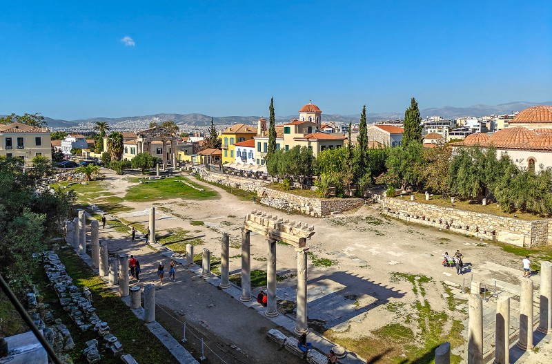 Best Time to Visit Plaka: Morning, Afternoon & Evening Walks - View over the Roman Agora in Athens with colonnades and gate, Plaka houses and cypress trees under a clear afternoon sky.