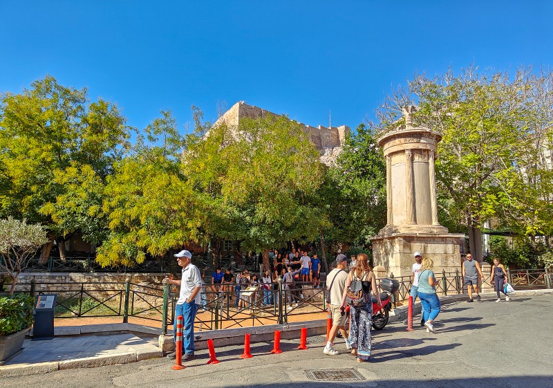 Monument of Lysicrates on Tripodon Street in Plaka, Athens, with the Acropolis and pedestrians in view.