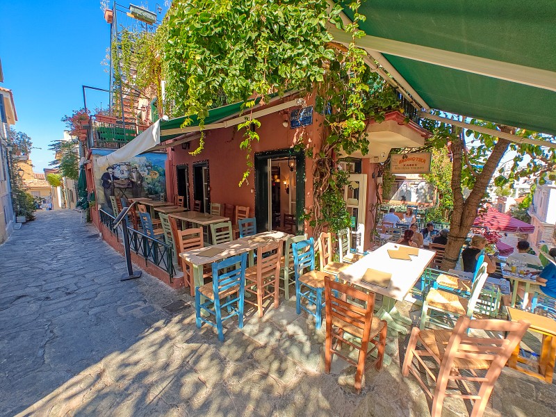 Restaurants in Athens - Traditional Greek taverna in Plaka, Athens, with colorful chairs, leafy vines, and outdoor tables along a cobblestone street