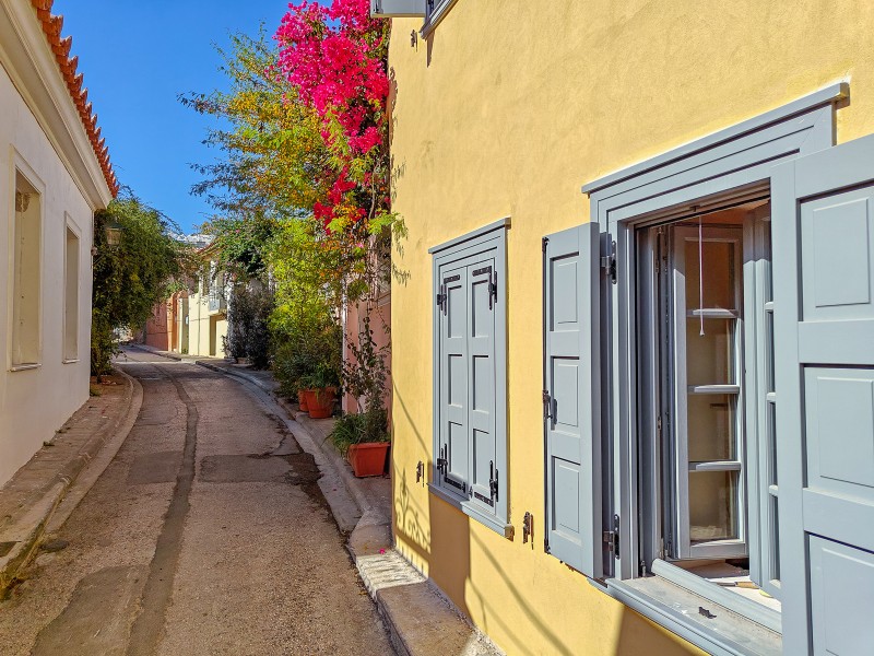 Quiet alley in Plaka, Athens, with pastel-colored houses, grey shutters, and blooming bougainvillea under the sun.
