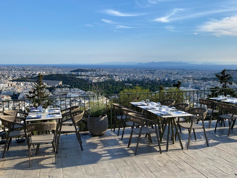 Outdoor restaurant terrace on Lycabettus Hill overlooking Athens, with panoramic city views stretching to the sea.


